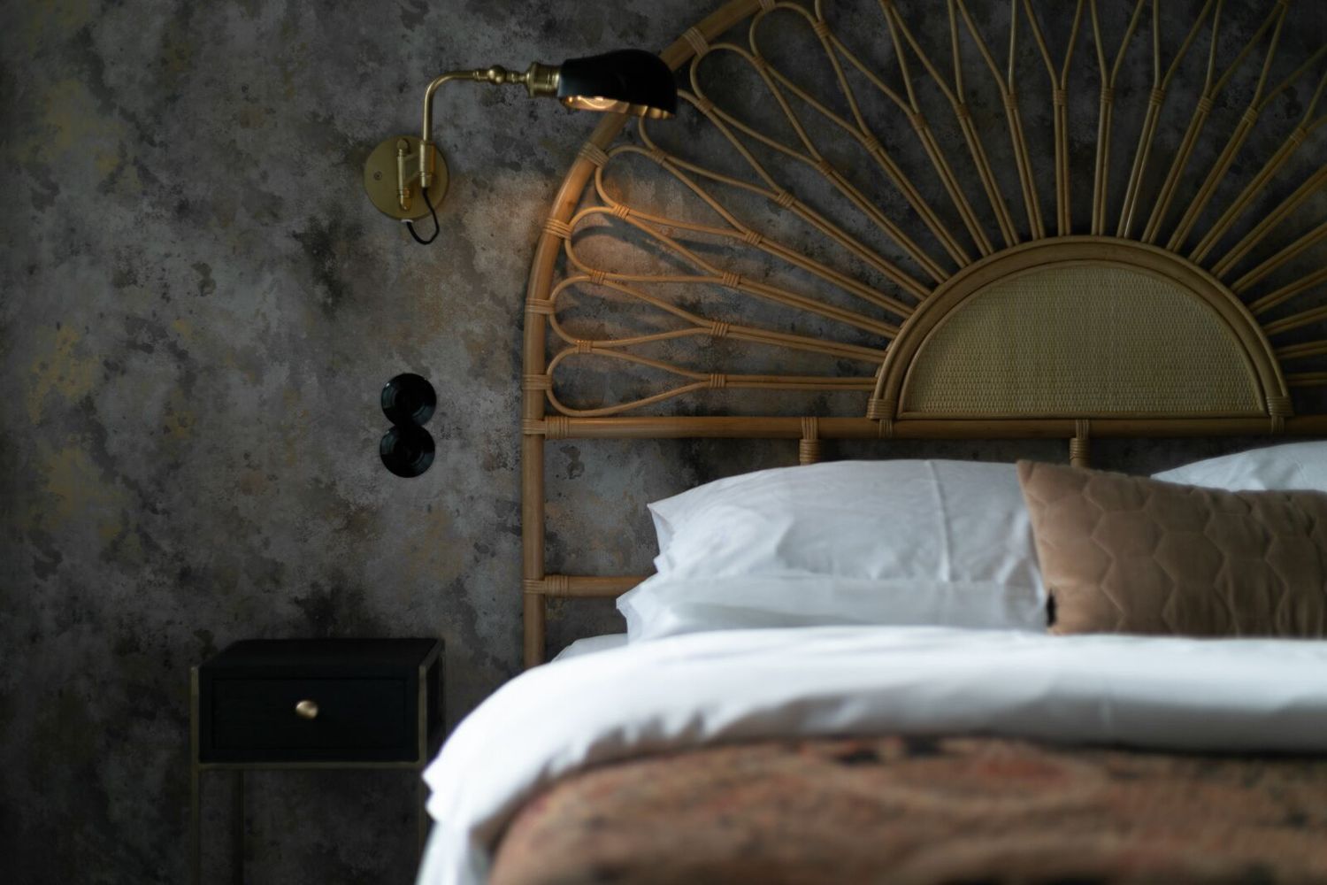 A cozy bedroom interior with a rattan headboard, white bedding, and brown accents against a textured gray wall.