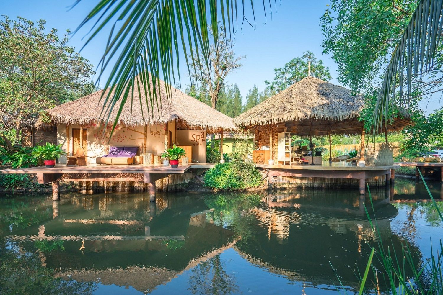 Tranquil overwater bungalows with thatched roofs reflect in calm lagoon waters surrounded by tropical palm trees.