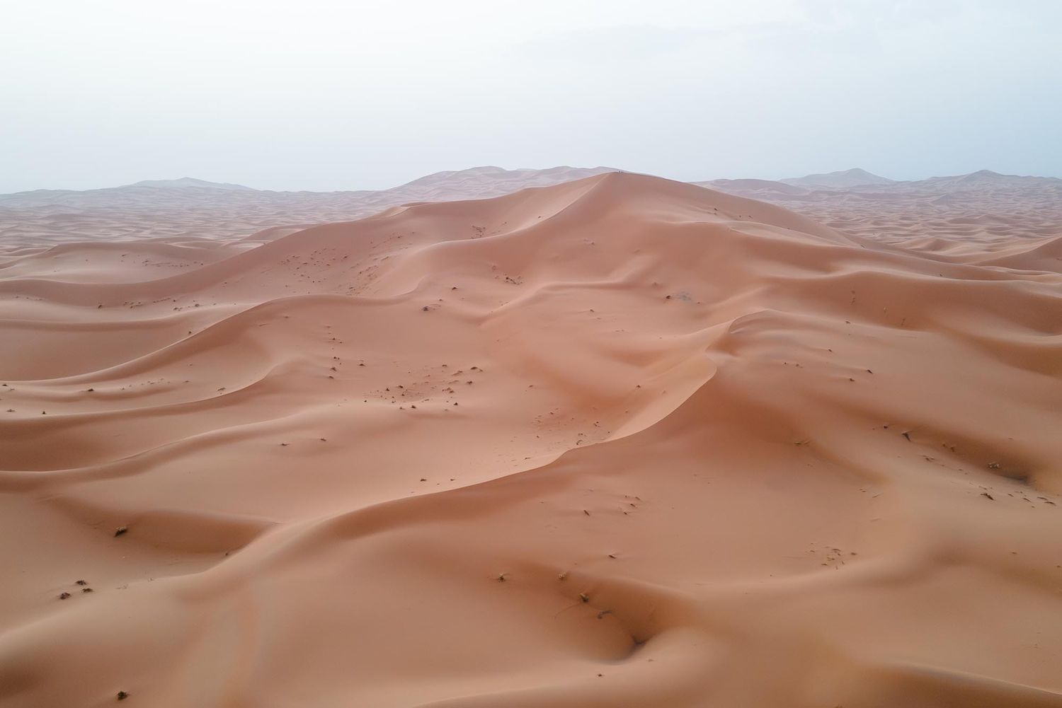 Aerial view of vast rolling sand dunes stretching across a desert landscape under a hazy sky.