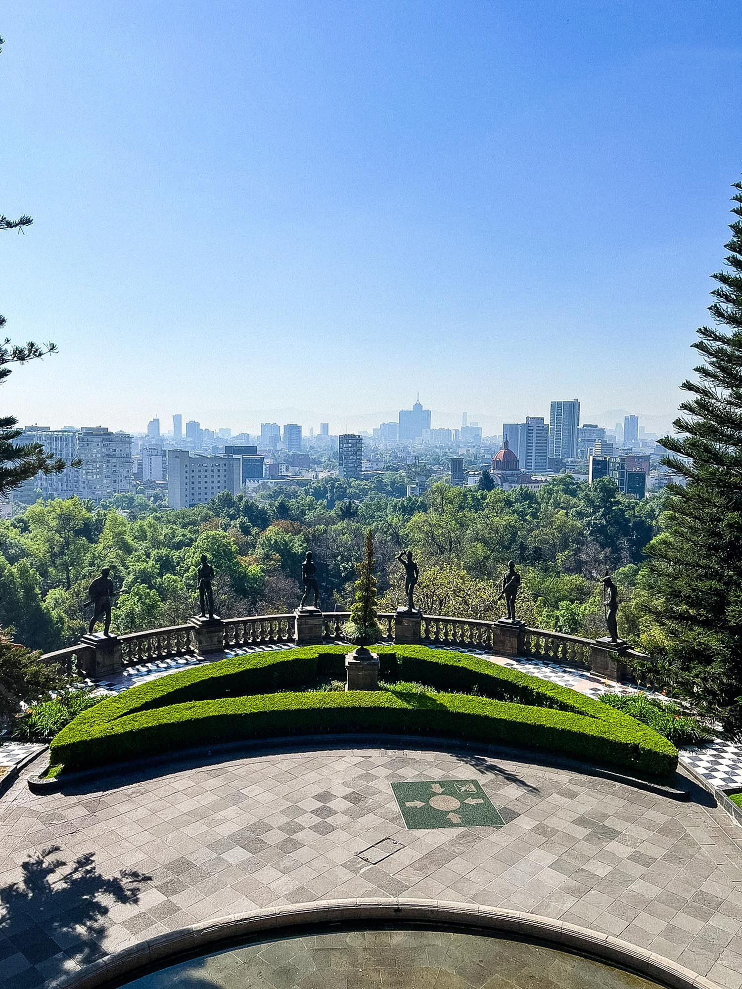 City skyline view from a curved stone balcony with manicured hedges and pine trees.