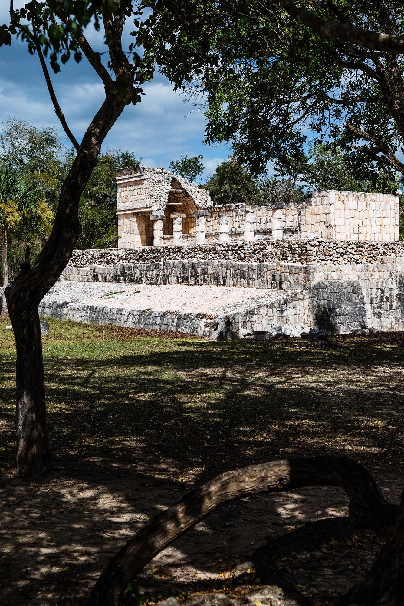 Ancient Mayan ruins with white stone walls and structures cast shadows under trees at an archaeological site in Mexico.