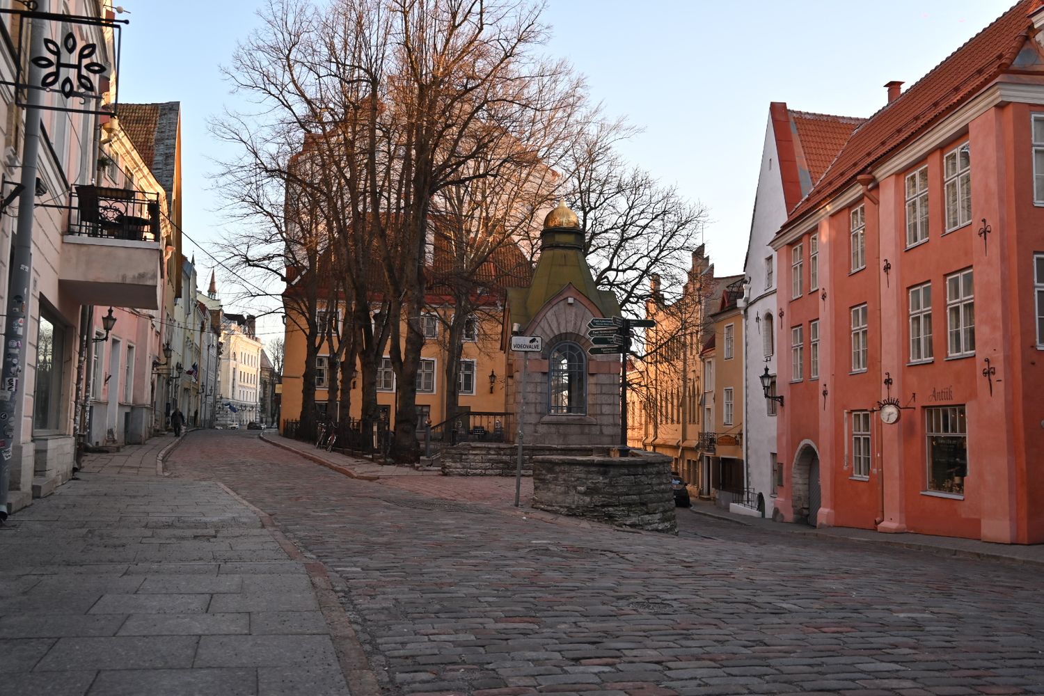 Cobblestone street lined with historic buildings and a stone cross in old town during sunset.