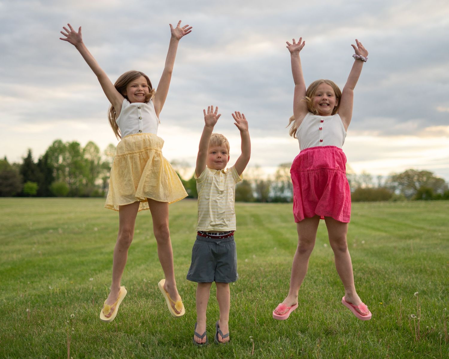Three joyful children jumping in a green field at sunset, two girls in colorful skirts raising arms high.