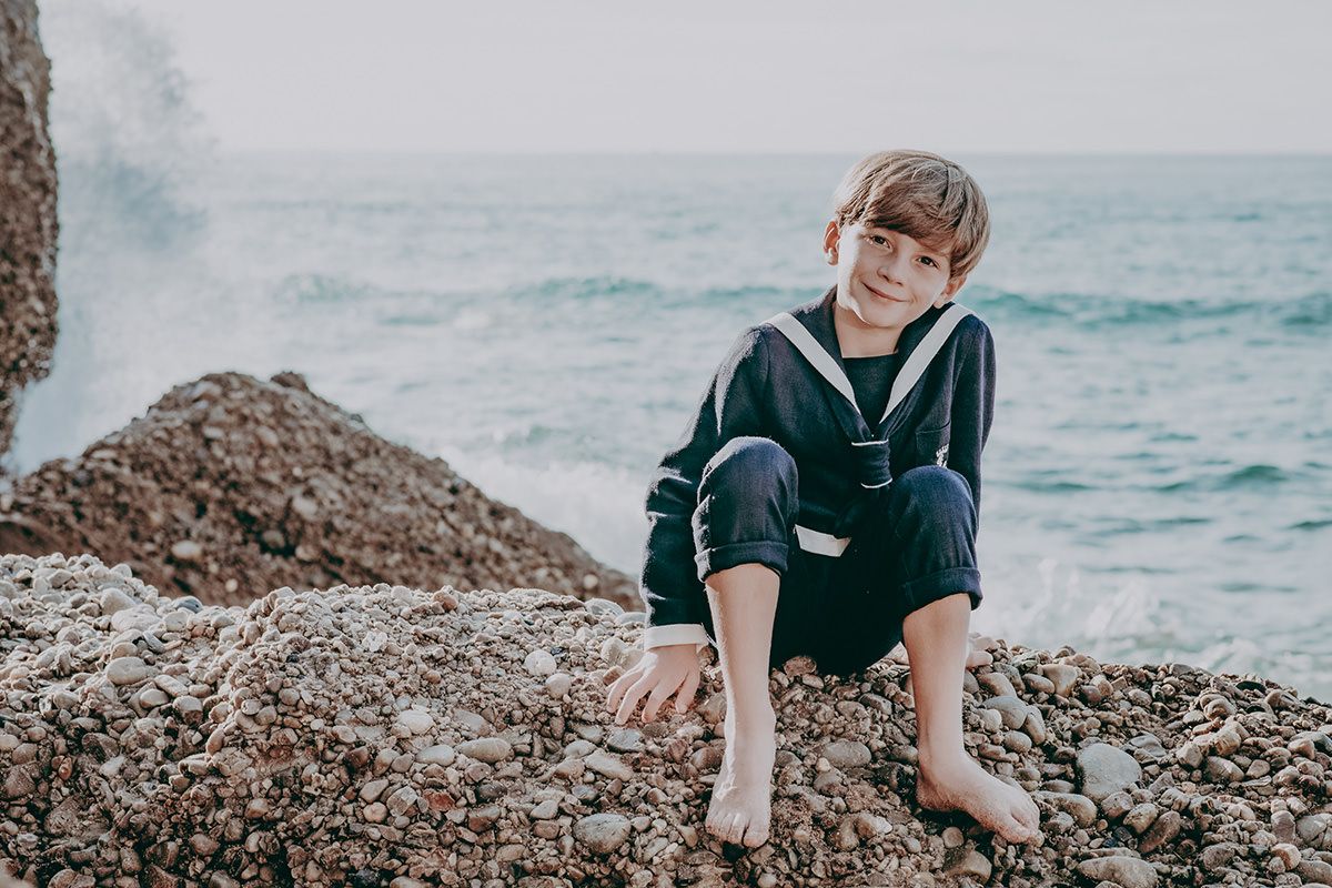 Niño de comunión sentado descalzo sobre las rocas en la playa con el mar Cantábrico de fondo en una playa de Gijón, Asturias.