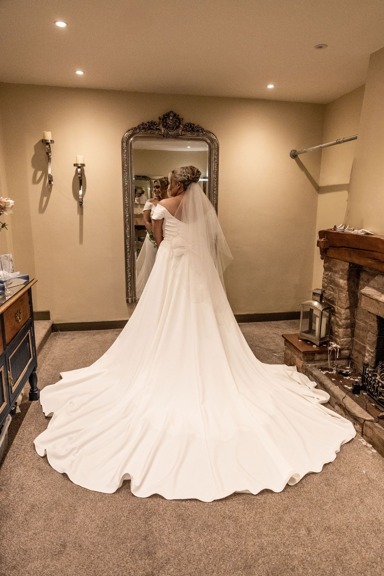 Bride in elegant white wedding gown with long train and veil, viewed from behind in mirror reflection.