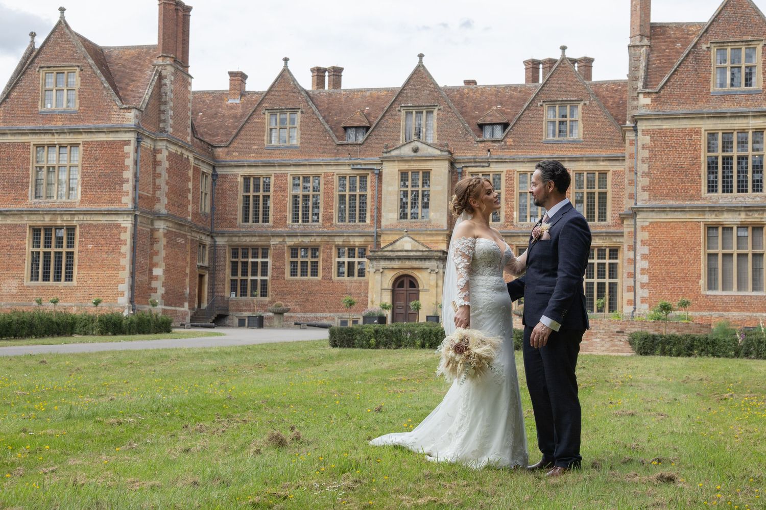 Bride and groom stand together on lawn before grand red brick English manor house wedding venue.