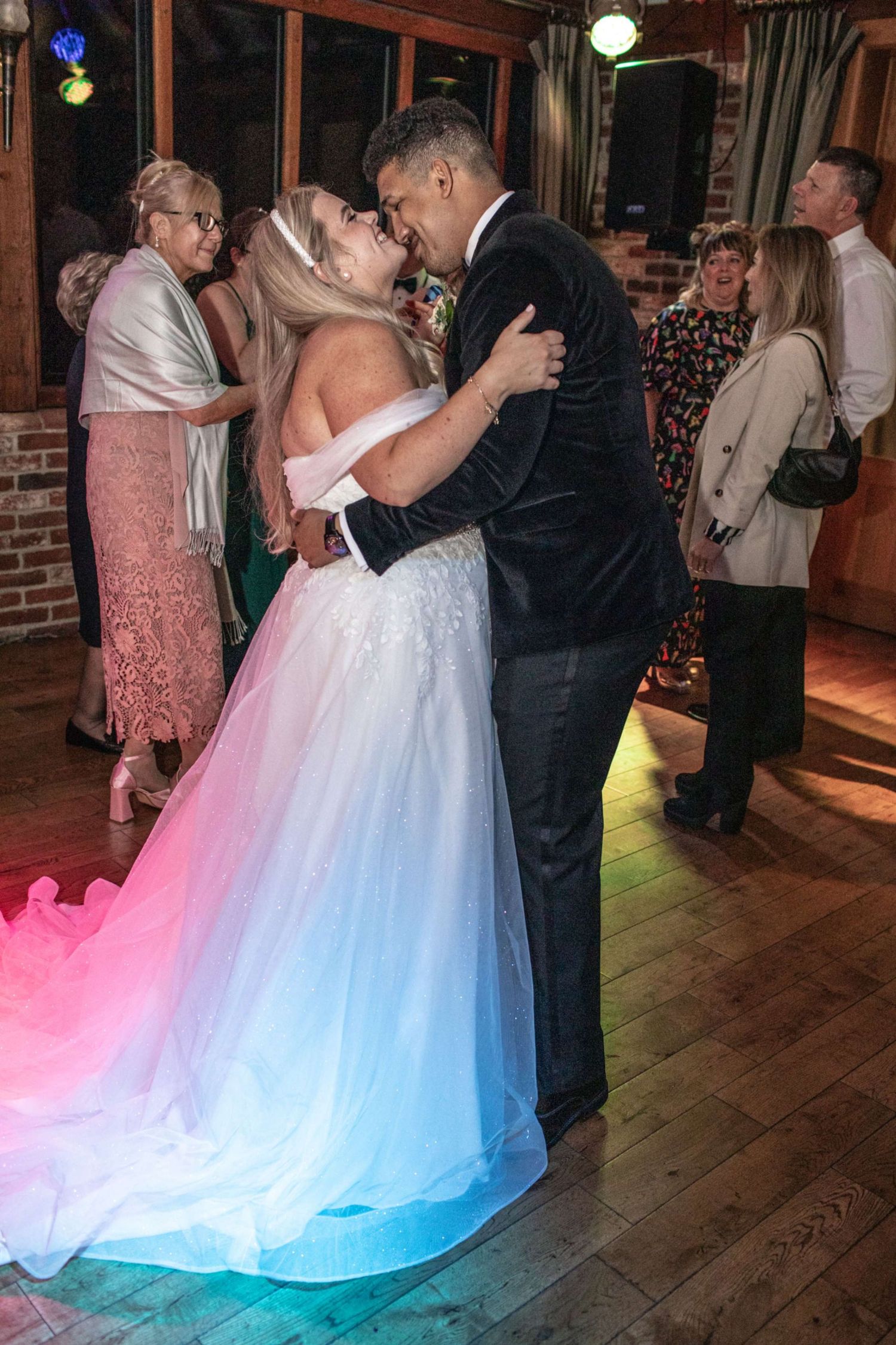 Bride and groom share first dance kiss in wedding venue with colorful lights, guests celebrating in background.