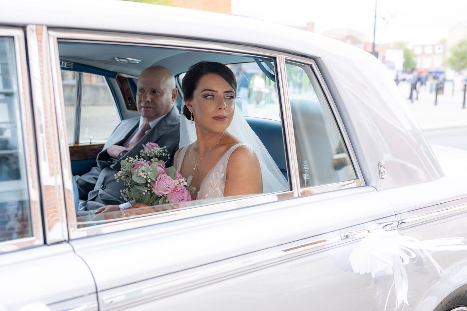 Bride holding pink bouquet sits in vintage white wedding car with groom, wearing elegant white gown.