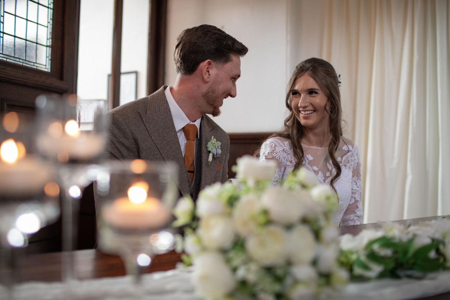 Bride and groom smiling at each other during wedding ceremony with candles and white floral arrangements in foreground.