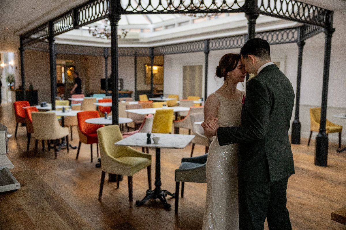 Bride and groom share a romantic kiss in an elegant restaurant with colorful chairs and ornate iron decor.