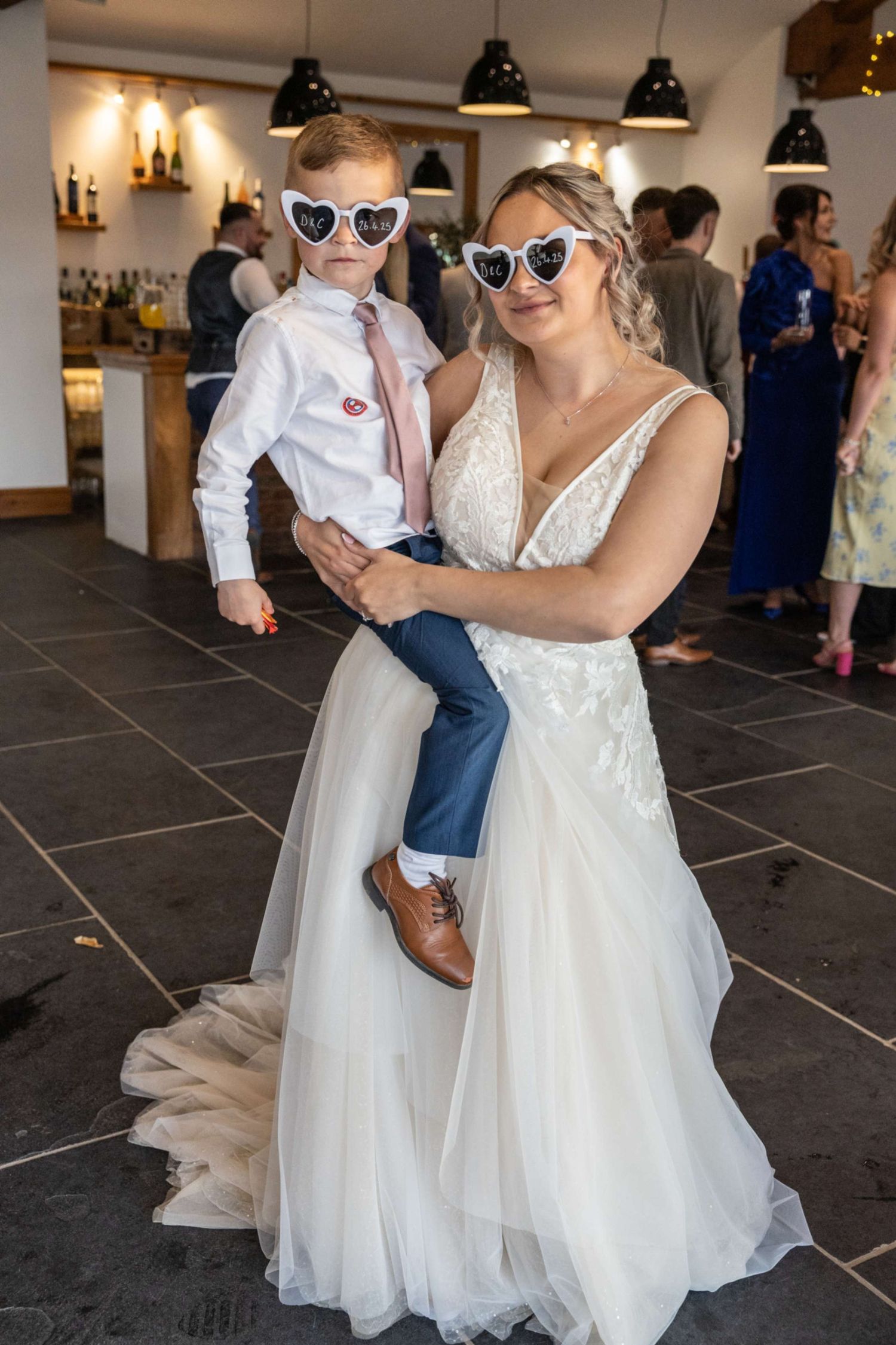 Bride in lace gown holds young boy, both wearing heart-shaped sunglasses at wedding reception.