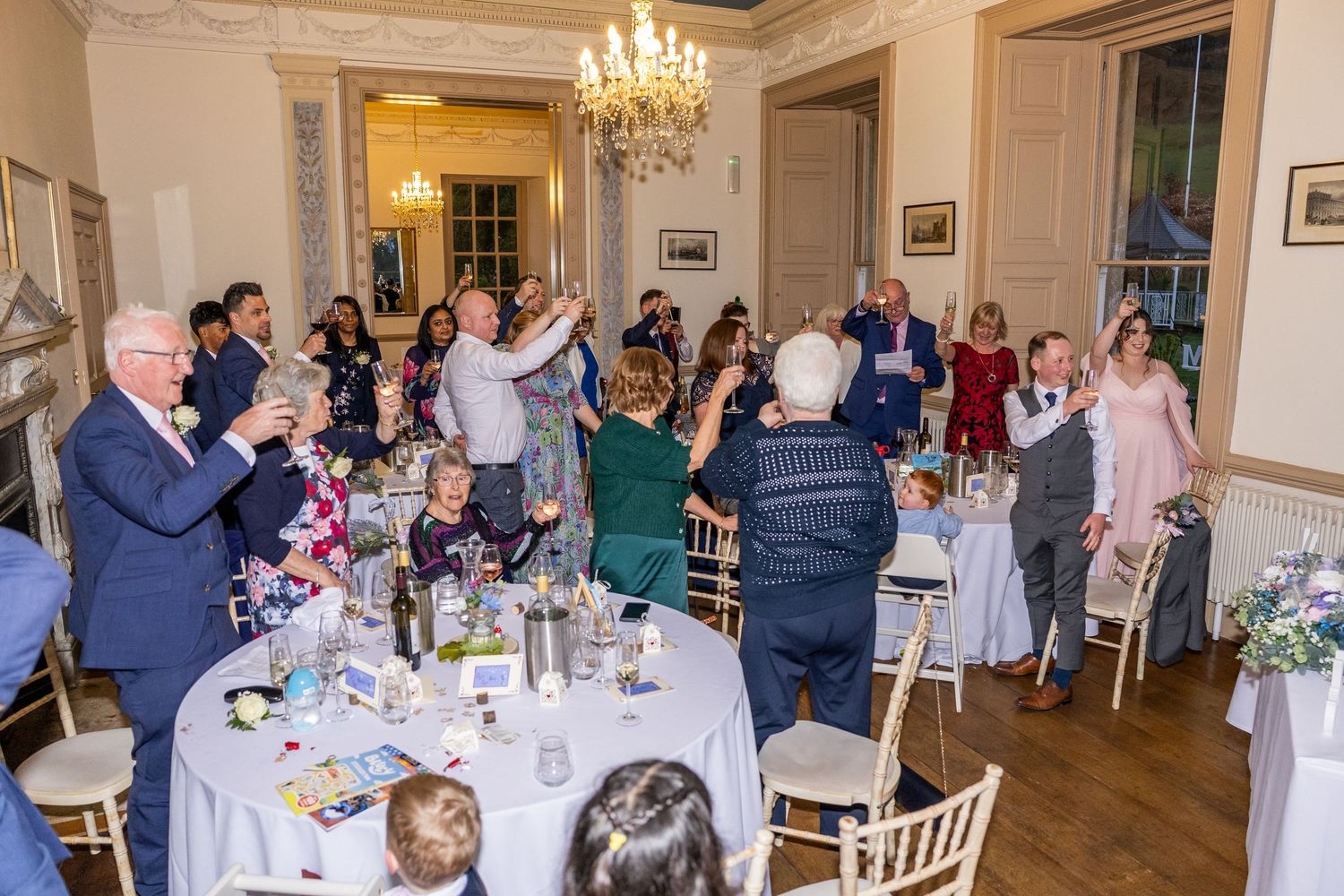 Wedding reception guests raising glasses in toast inside elegant chandelier-lit dining room with round tables.