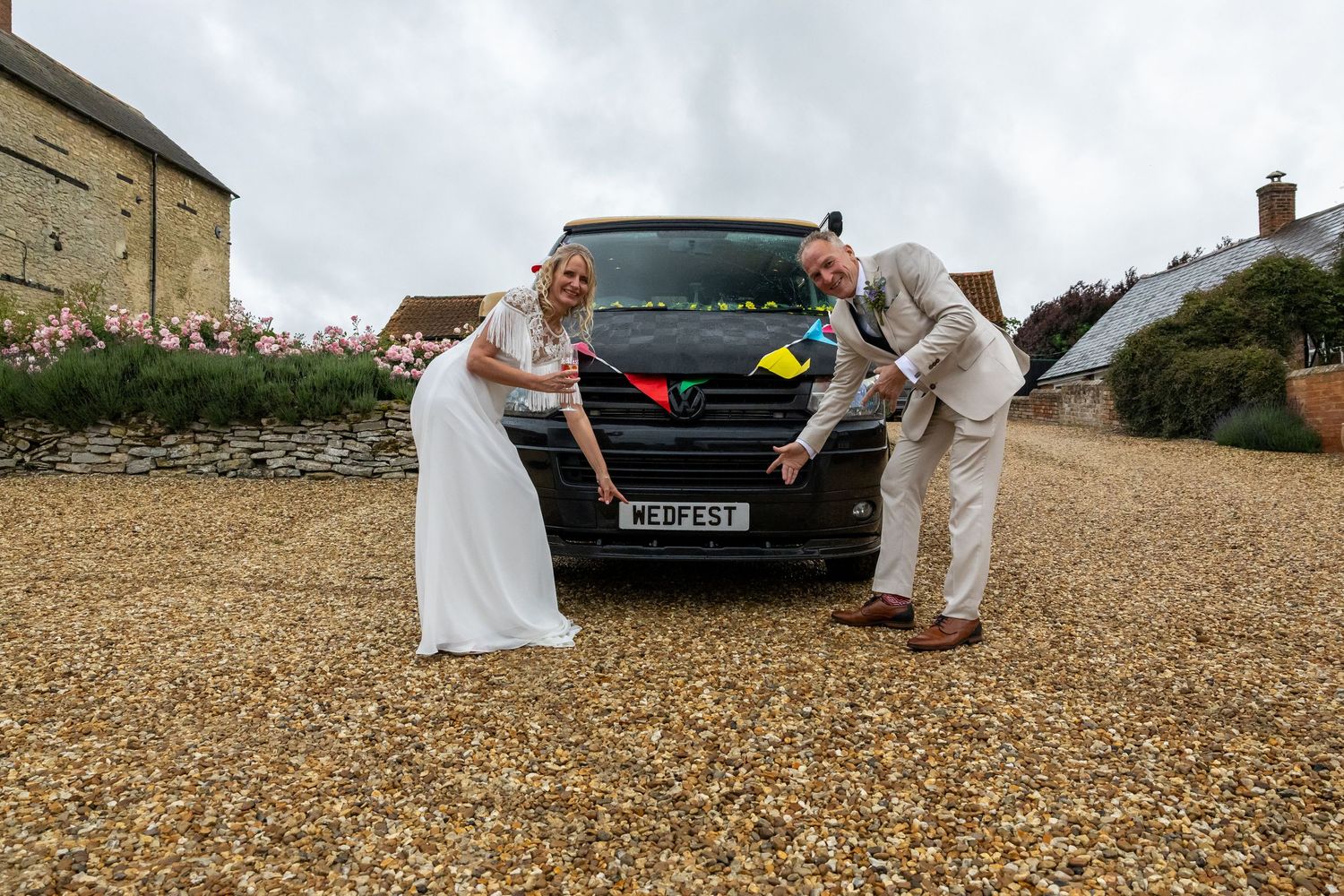 Bride and groom posing with a black VW van decorated with colorful bunting on a gravel driveway at a countryside wedding.
