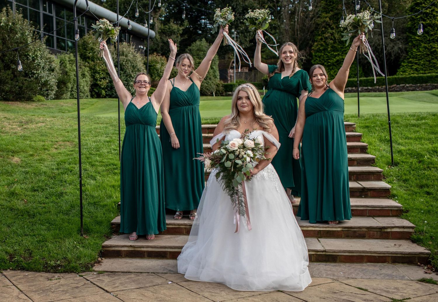 Bride in white gown holds bouquet with four bridesmaids in emerald green dresses celebrating on stone steps outdoors.