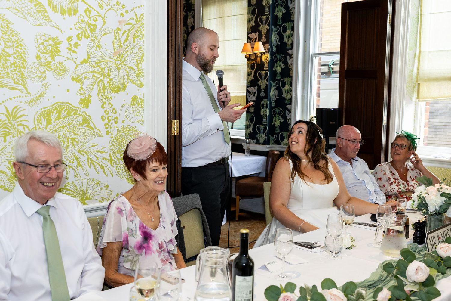 Man giving a speech at a wedding reception dinner, guests smiling at decorated tables in an elegant venue.