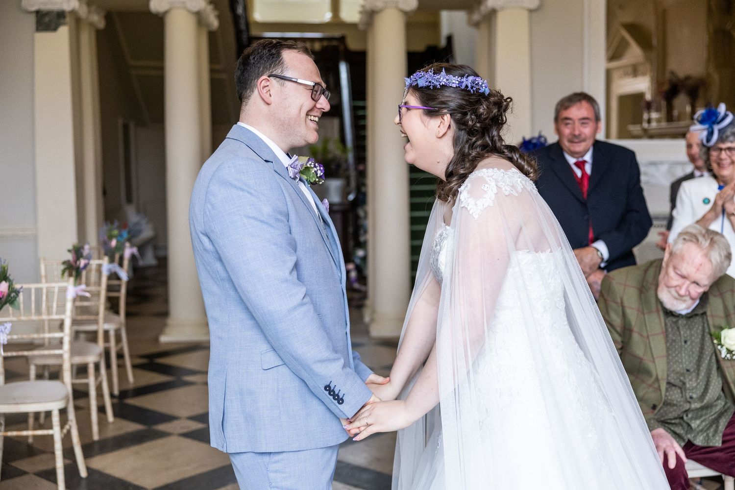 A joyful bride and groom hold hands and laugh during their wedding ceremony in an elegant venue with columns.