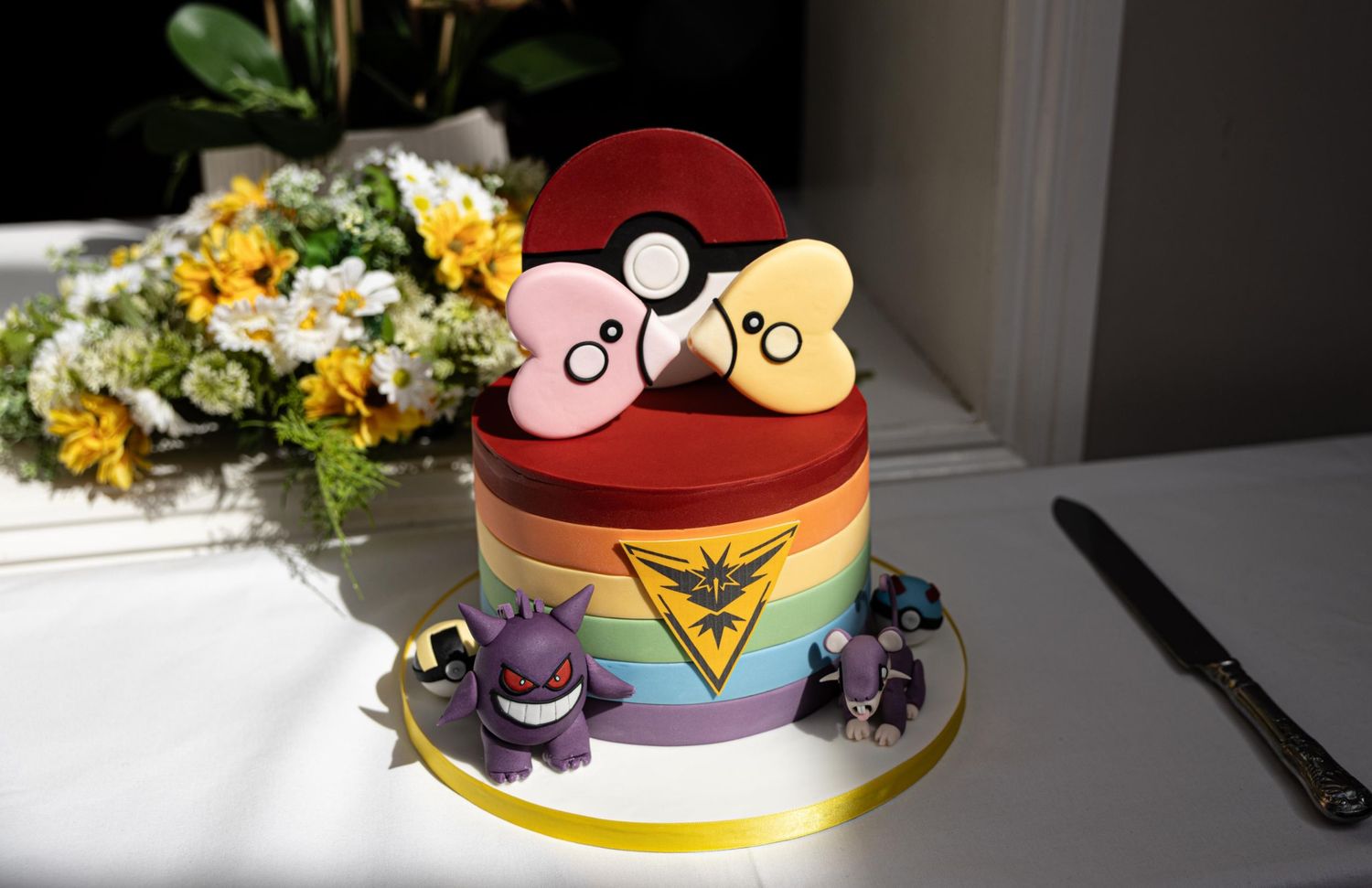 Rainbow Pokemon wedding cake with Pokeball topper, Gengar and other Pokemon figurines, beside flowers.