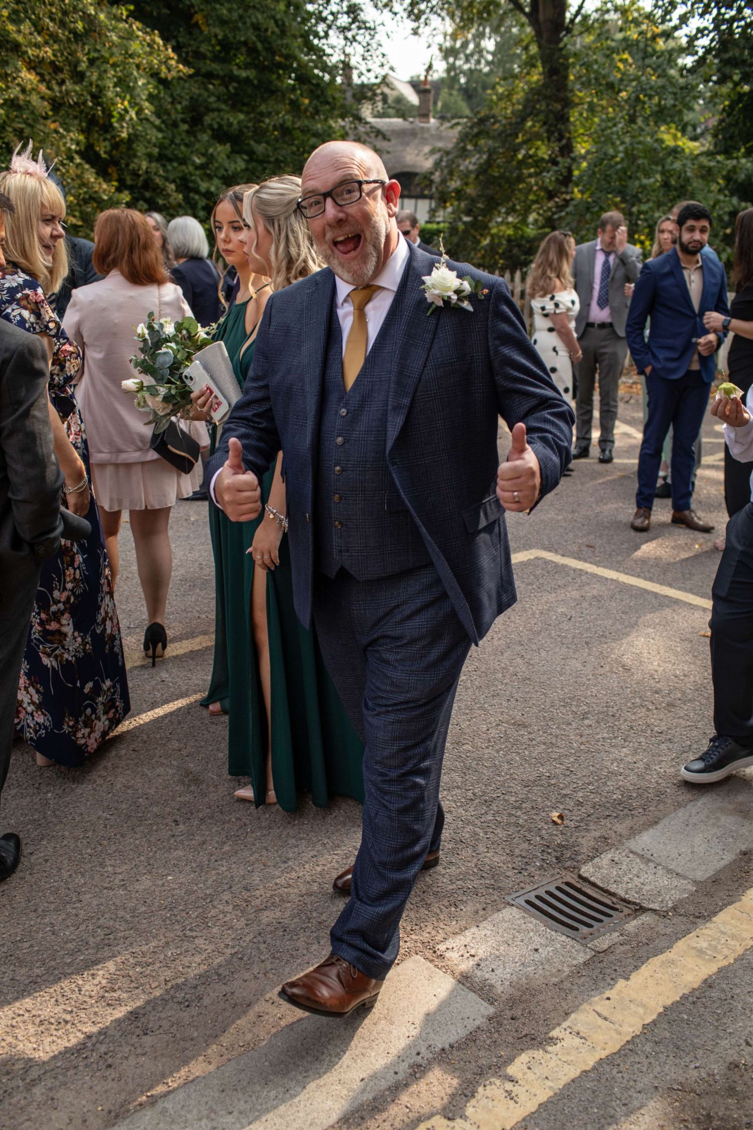 Smiling bald man in navy suit giving thumbs up at outdoor wedding celebration with guests in background.