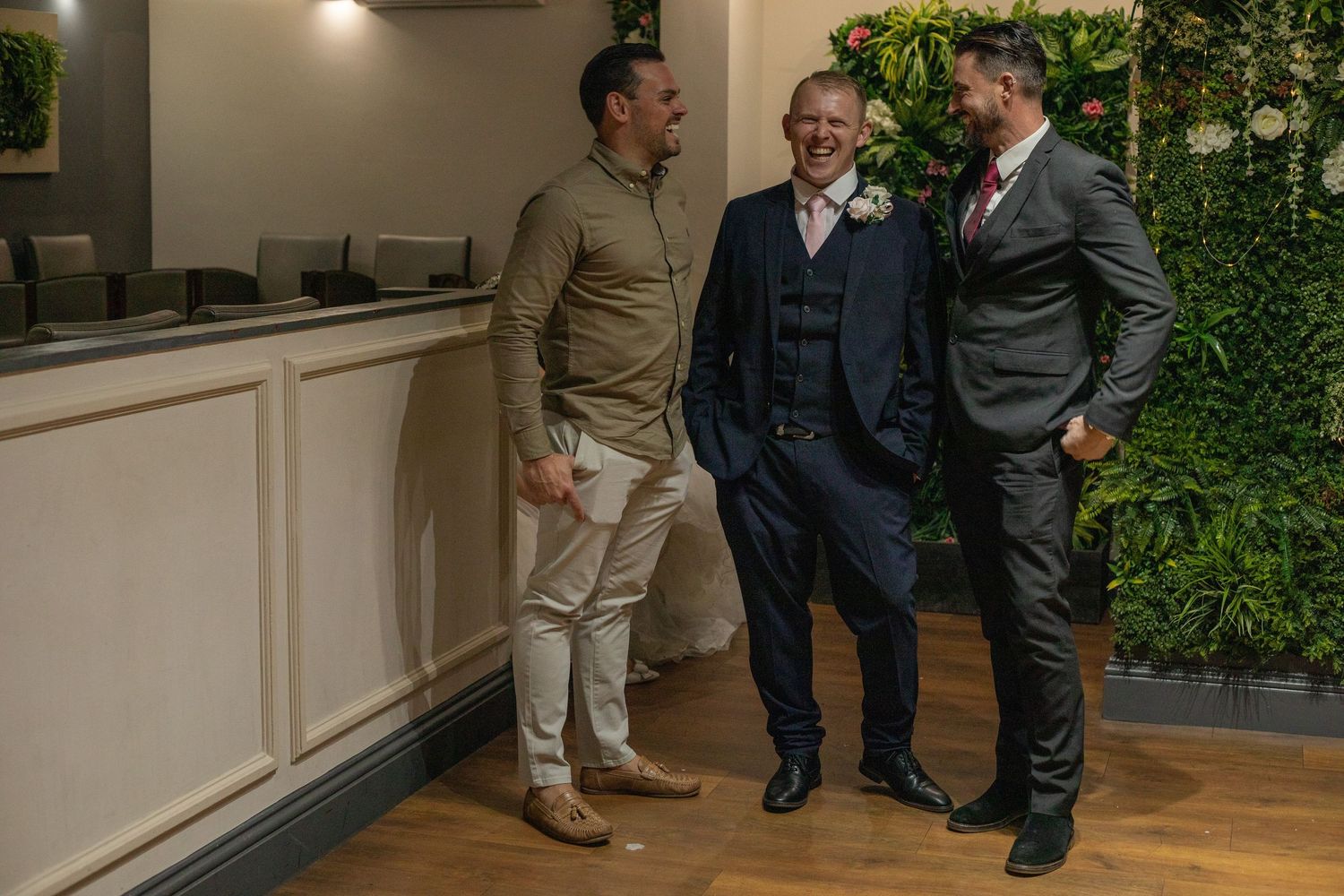 Three men laughing together at a wedding venue with a green living wall backdrop and wooden flooring.