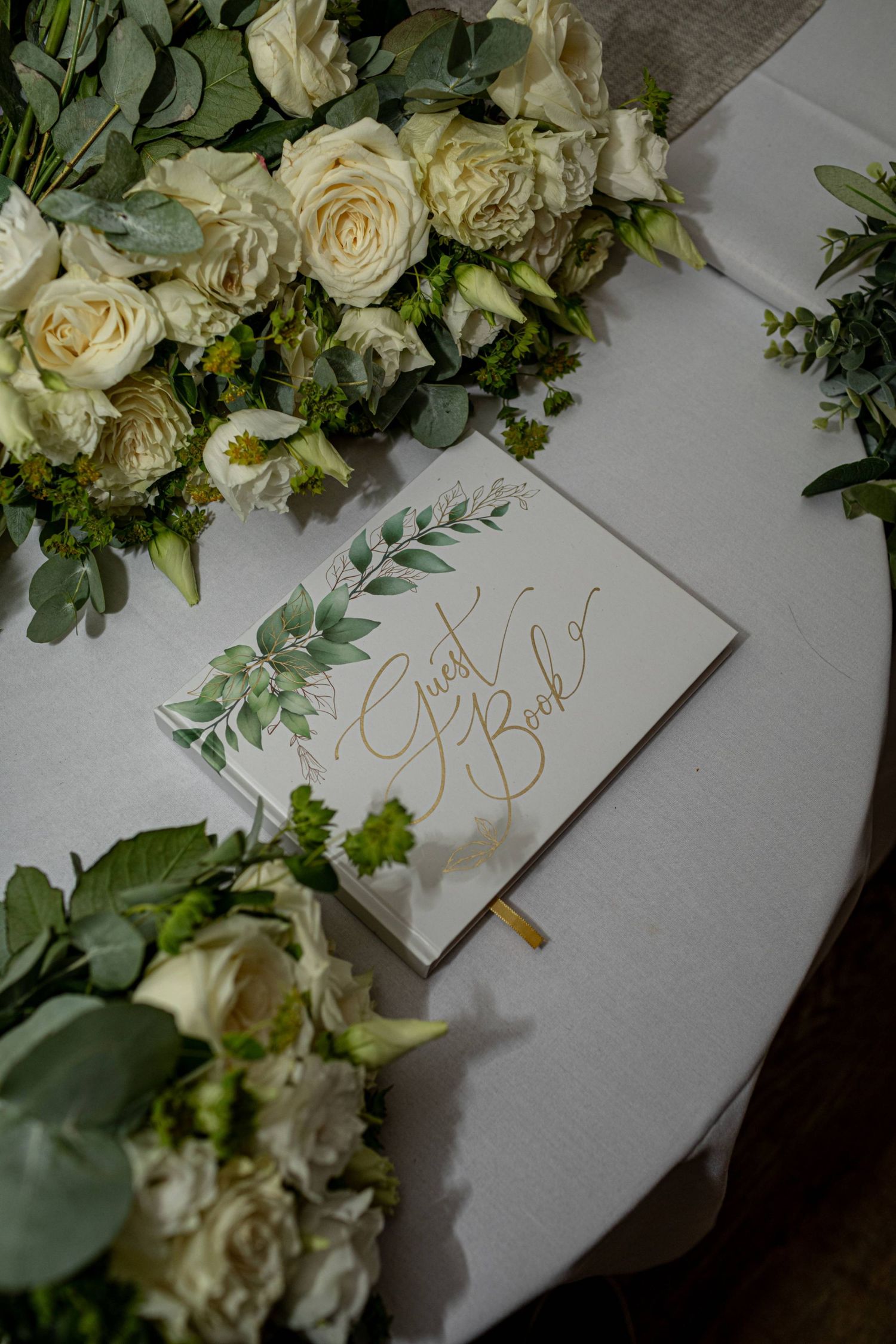 Wedding guest book with gold calligraphy surrounded by elegant white roses and greenery on a white tablecloth.