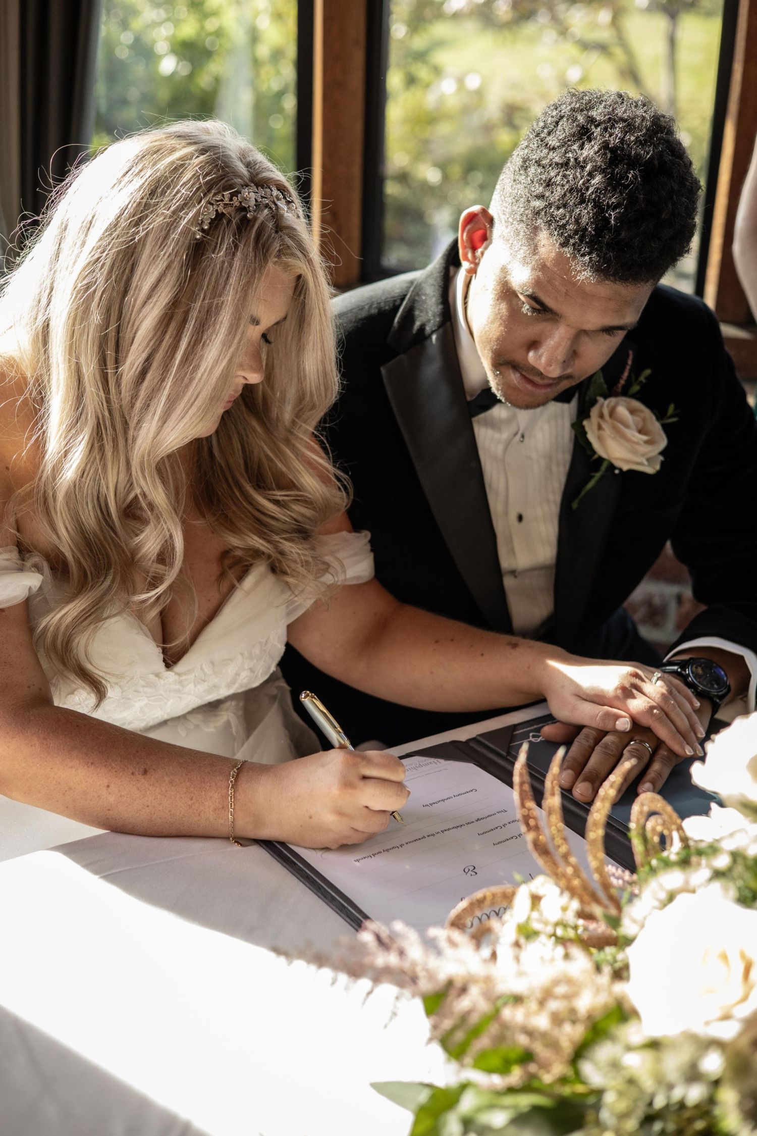 Bride signs wedding certificate as groom watches, both dressed in formal wedding attire at decorated table.