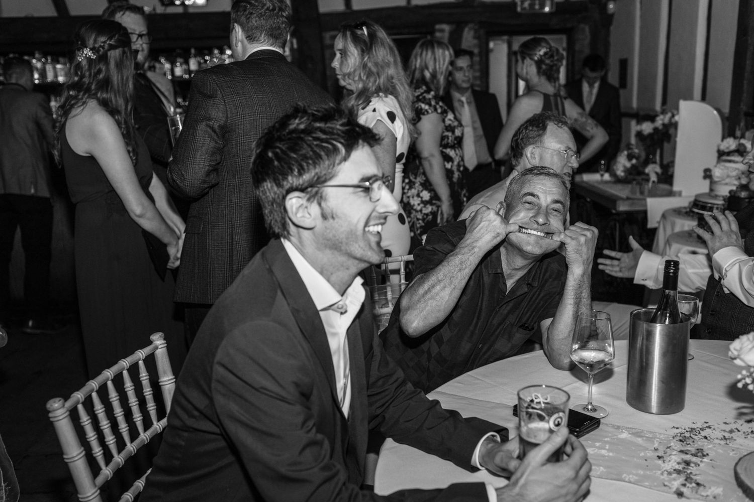 Black and white photo of two men laughing at a wedding reception dinner table with guests dancing in background.