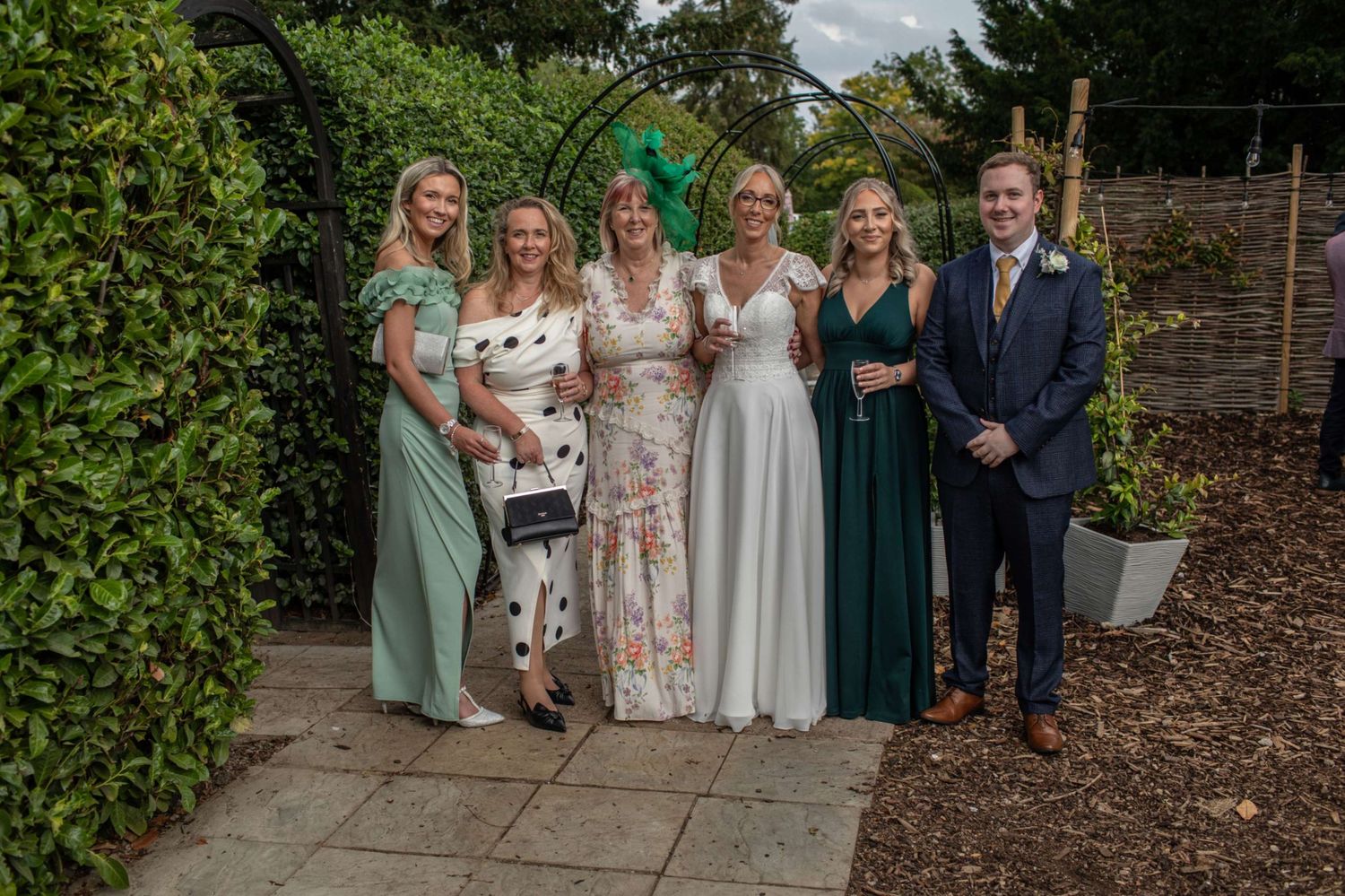 Six elegantly dressed wedding guests pose in a garden, with the bride in white and bridesmaids in green gowns.