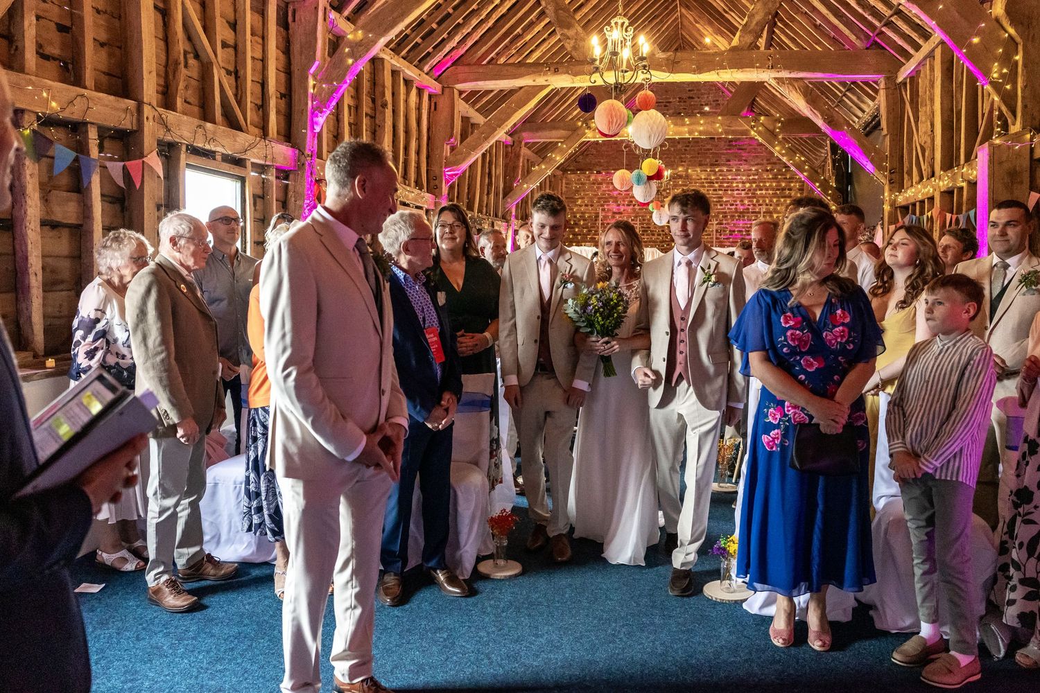 Rustic barn wedding ceremony with guests standing as bride and groom exchange vows under warm pink lights.