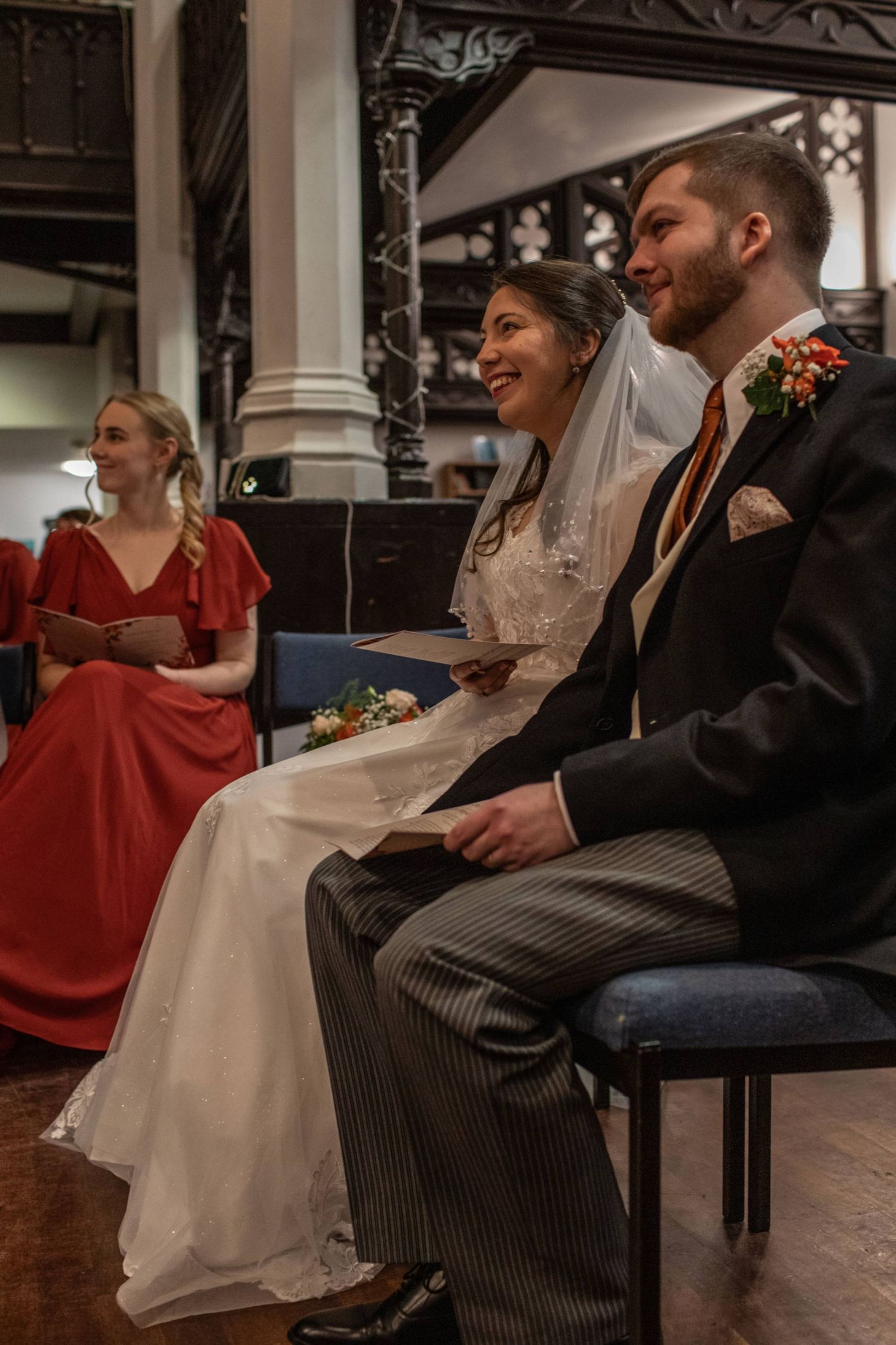 Bride and groom seated at wedding ceremony in church, smiling joyfully, with bridesmaid in red dress in background.