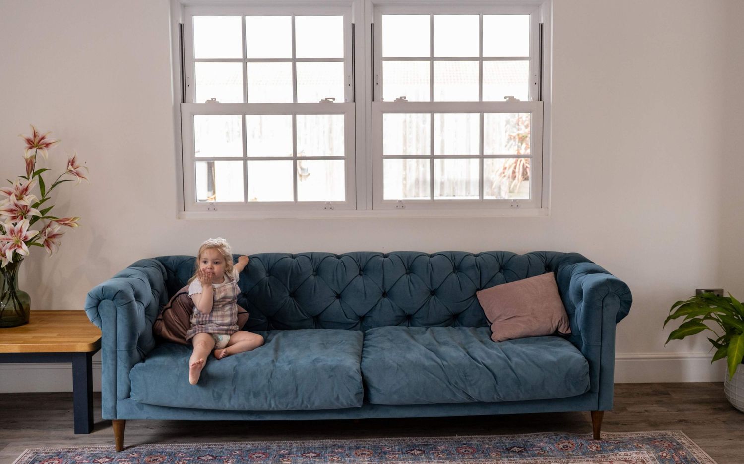 Baby sitting on a tufted blue velvet Chesterfield sofa in a bright white living room.