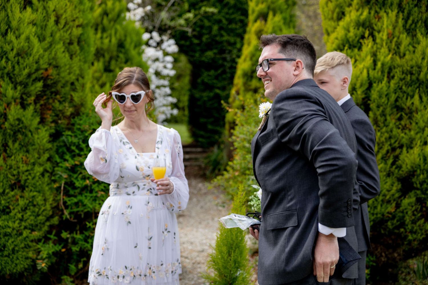 Wedding guests socializing outdoors, woman in white floral dress adjusting sunglasses while holding a drink.