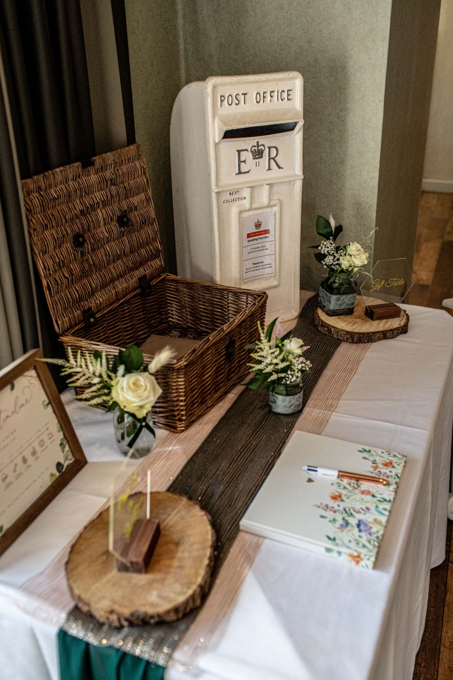 Wedding gift table with white Post Office letter box, wicker hamper, guest book, and floral arrangements on wood slice decor.