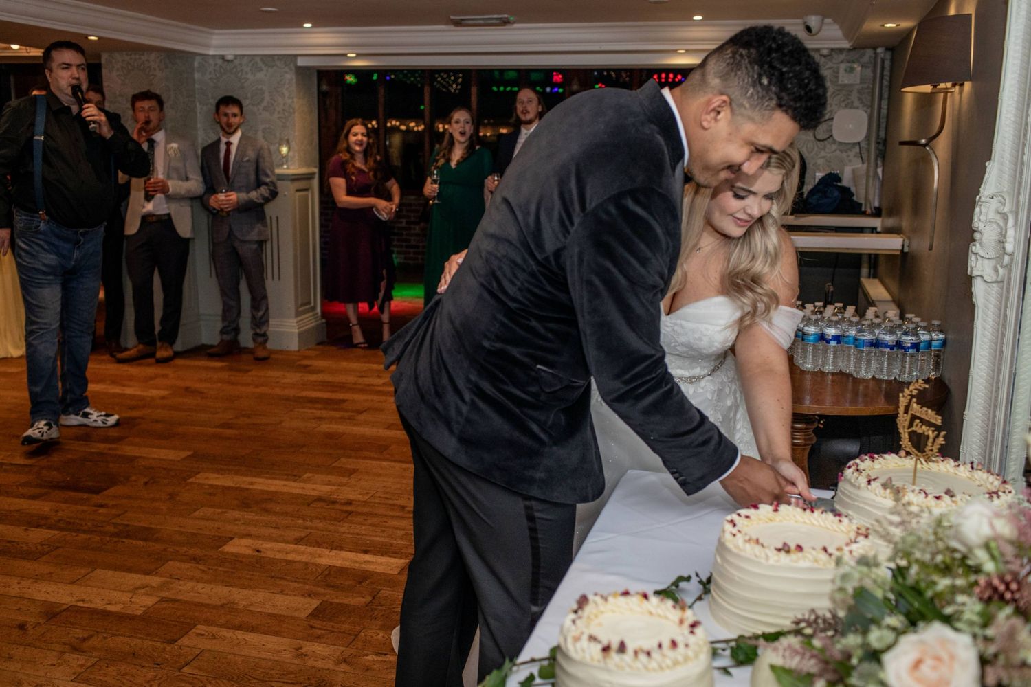 Bride and groom cutting their white wedding cake together at a romantic indoor reception with guests watching.