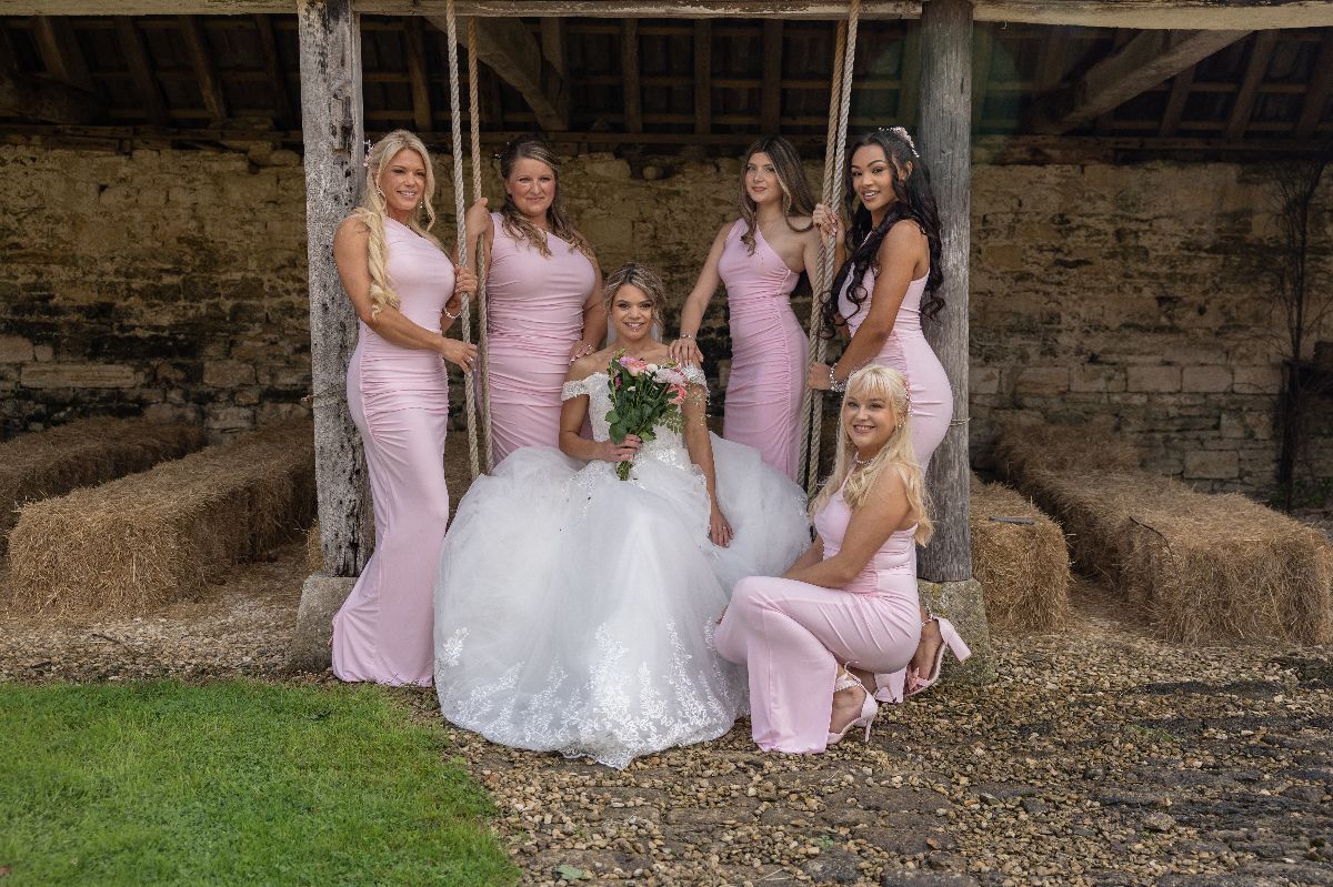 Bride in white gown poses with five bridesmaids in pink dresses inside a rustic barn with hay bales.