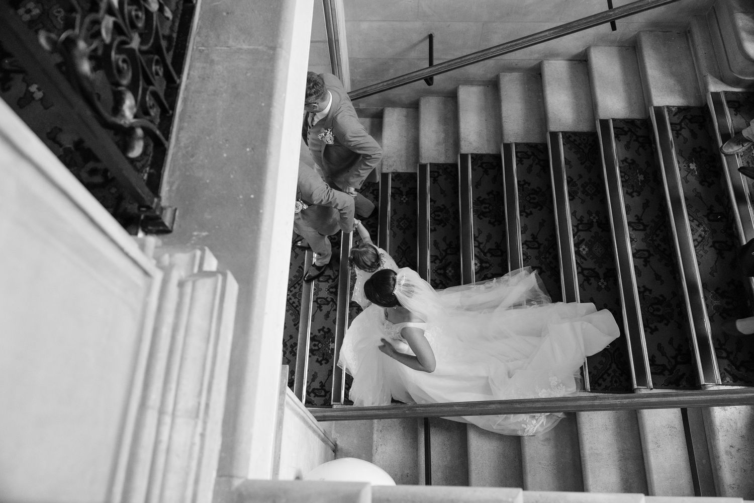 A black and white photograph of a bride in a flowing wedding dress ascending an elegant staircase.