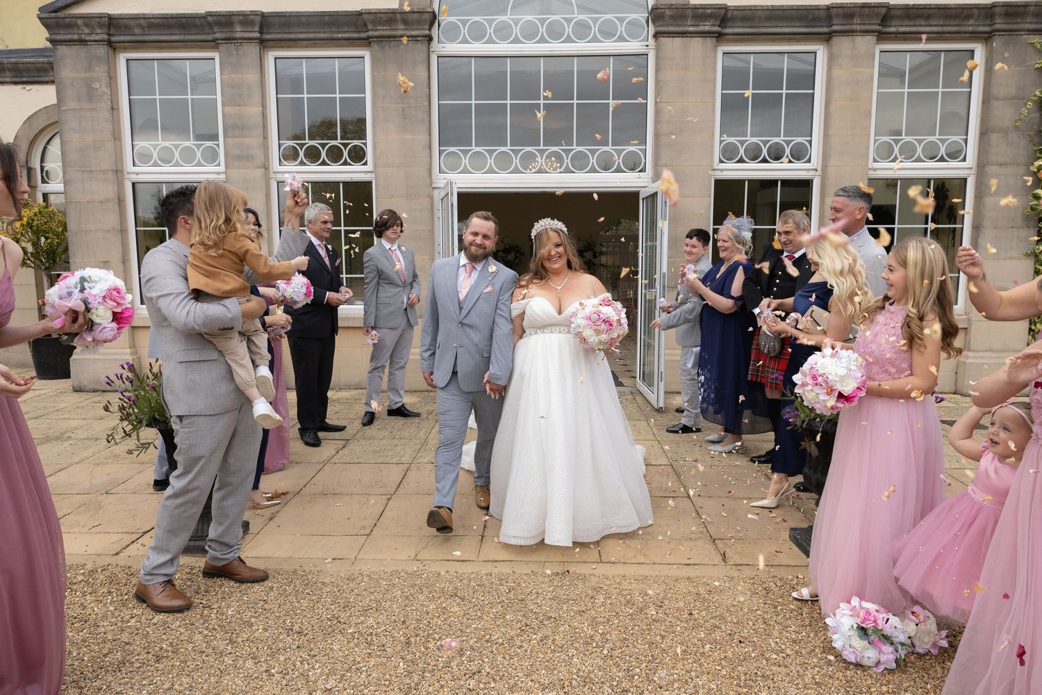 Bride and groom walk through confetti shower with wedding guests in pink dresses outside elegant venue.