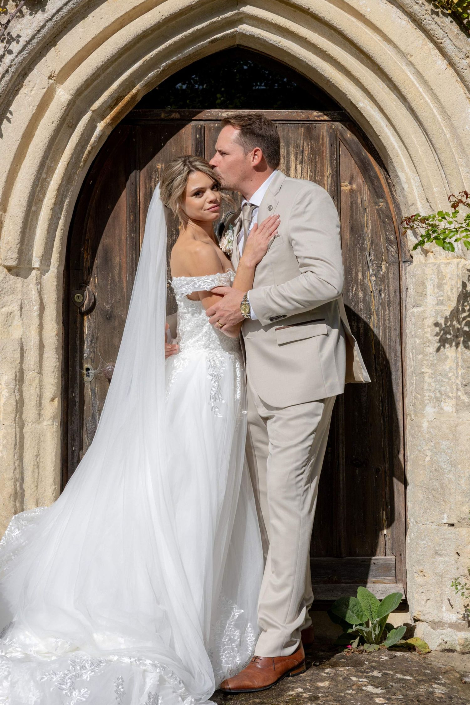 Bride and groom sharing a kiss in front of a rustic stone church doorway, bride in white gown with veil.