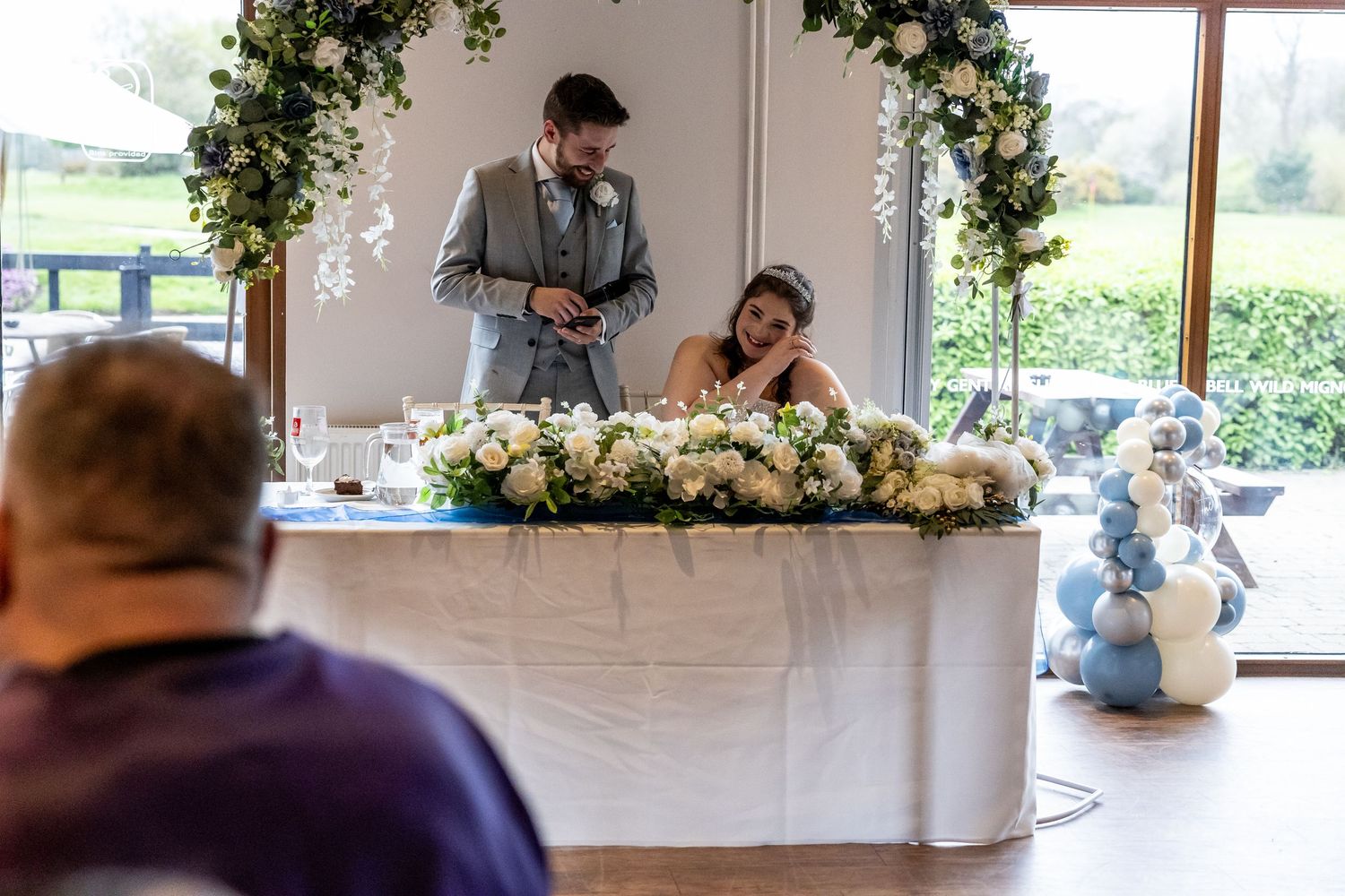 Bride and groom at floral wedding reception head table with white flowers and balloon decorations.