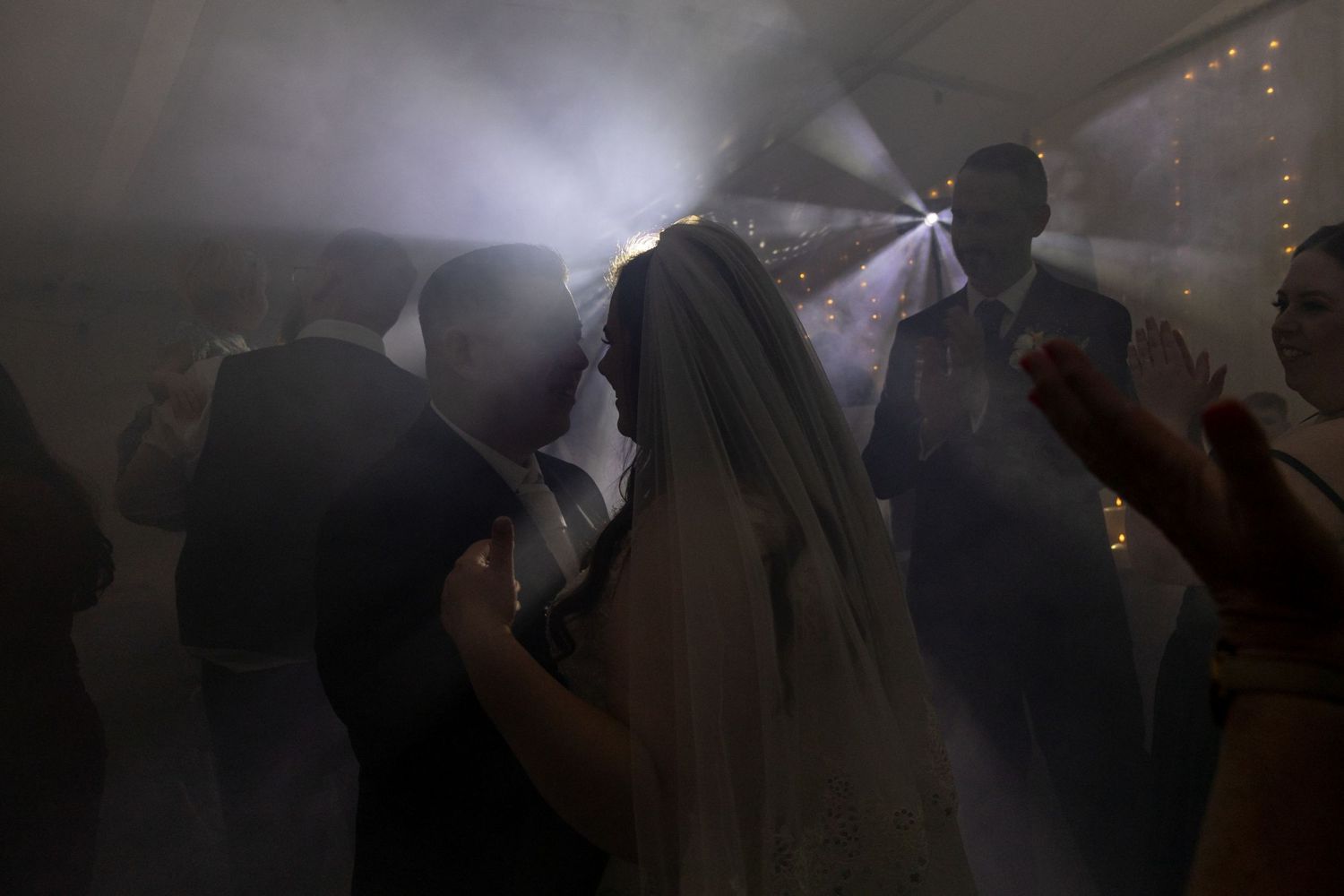 Bride in veil dancing with guests in smoky, atmospheric wedding reception with dramatic lighting.