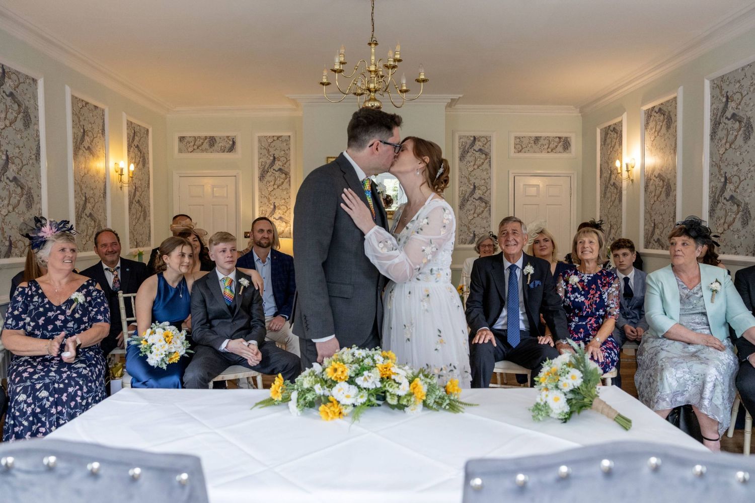 Bride and groom share first kiss at elegant wedding ceremony with seated guests and yellow floral arrangements.