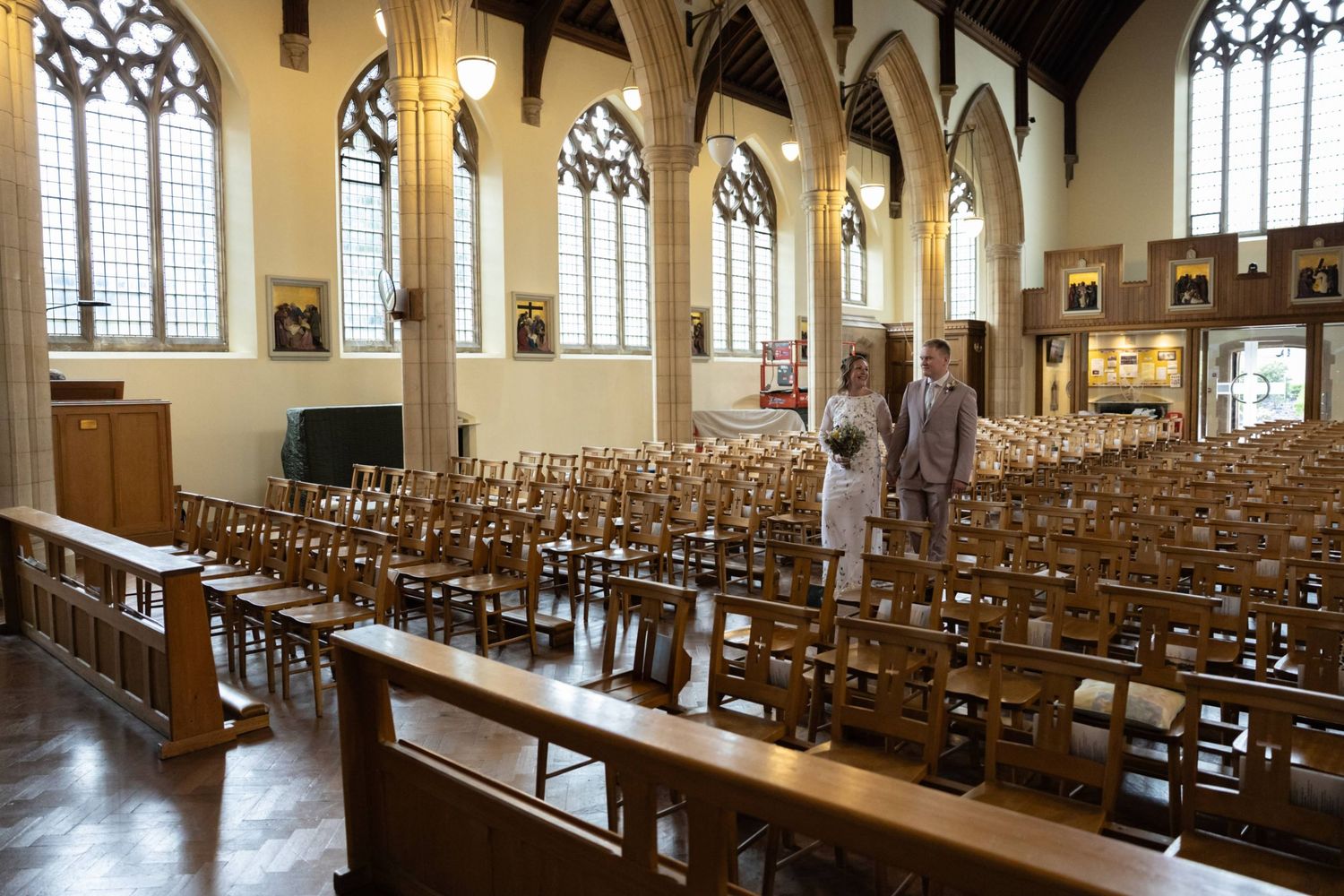 Two people stand in an empty historic church hall with rows of wooden chairs and Gothic stained glass windows.