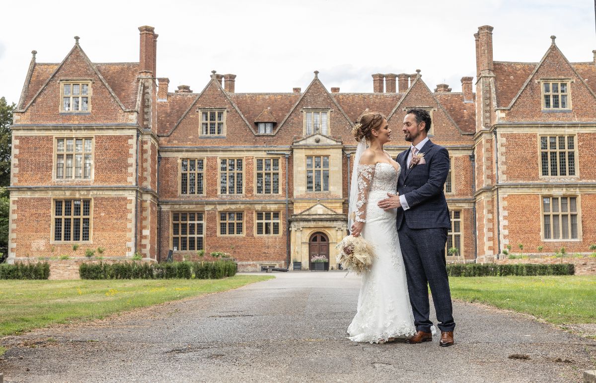 Bride and groom stand together on a path before a grand red-brick English manor house on their wedding day.