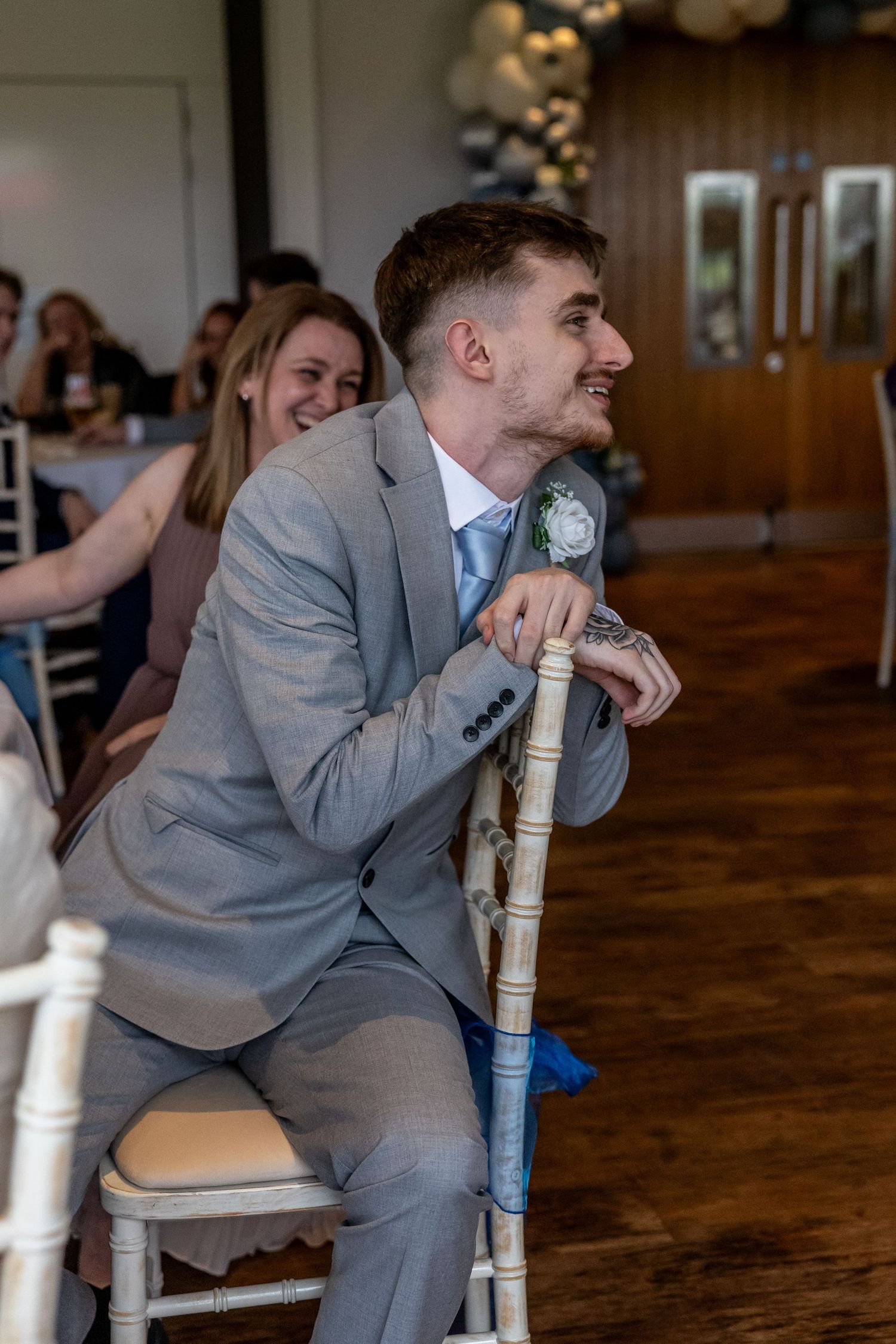 A young man in a gray suit with a boutonniere sits on a white chair at a wedding reception, smiling.