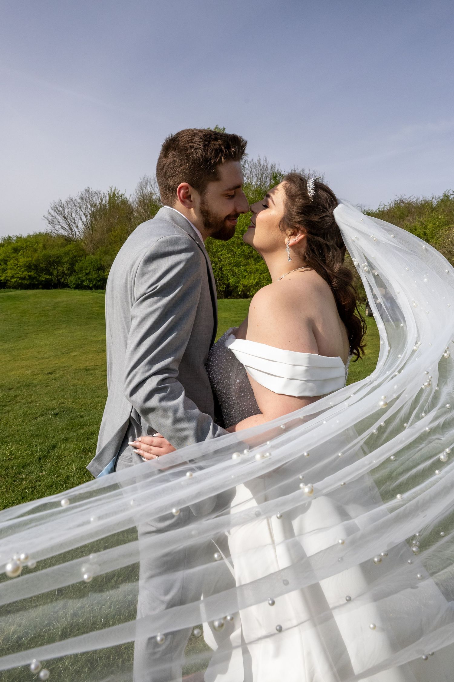 Bride and groom sharing a romantic almost-kiss outdoors, with her pearl-adorned veil flowing in the breeze.