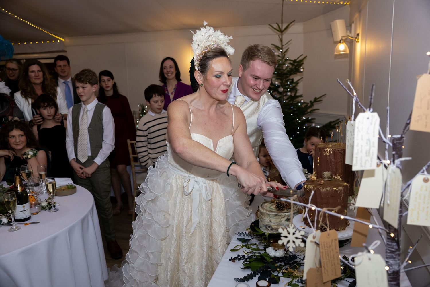 Wedding reception guests gather around tables decorated with white linens and centerpieces in an elegant indoor venue.