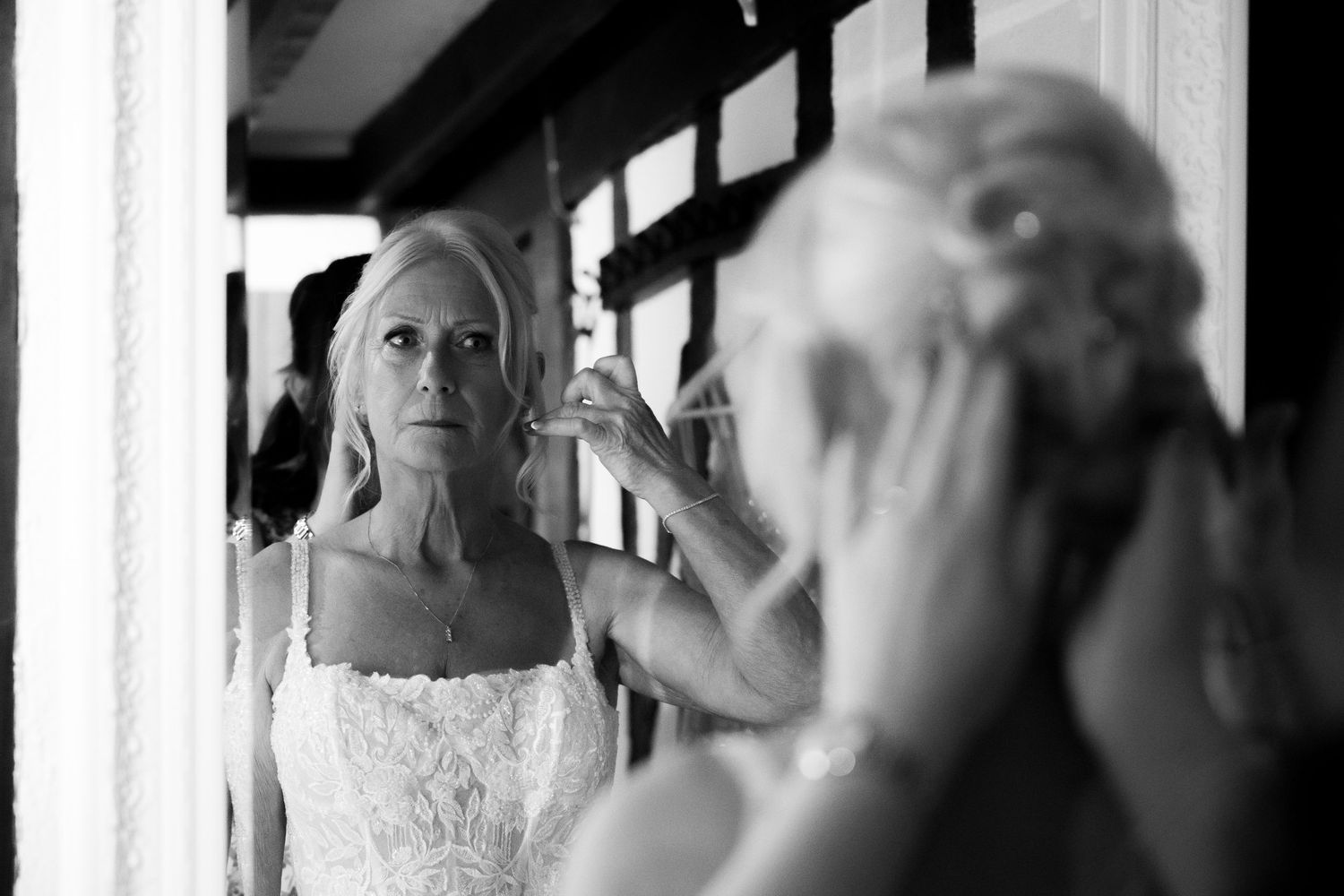 Black and white photo of a bride adjusting her earring while looking in a mirror on her wedding day.