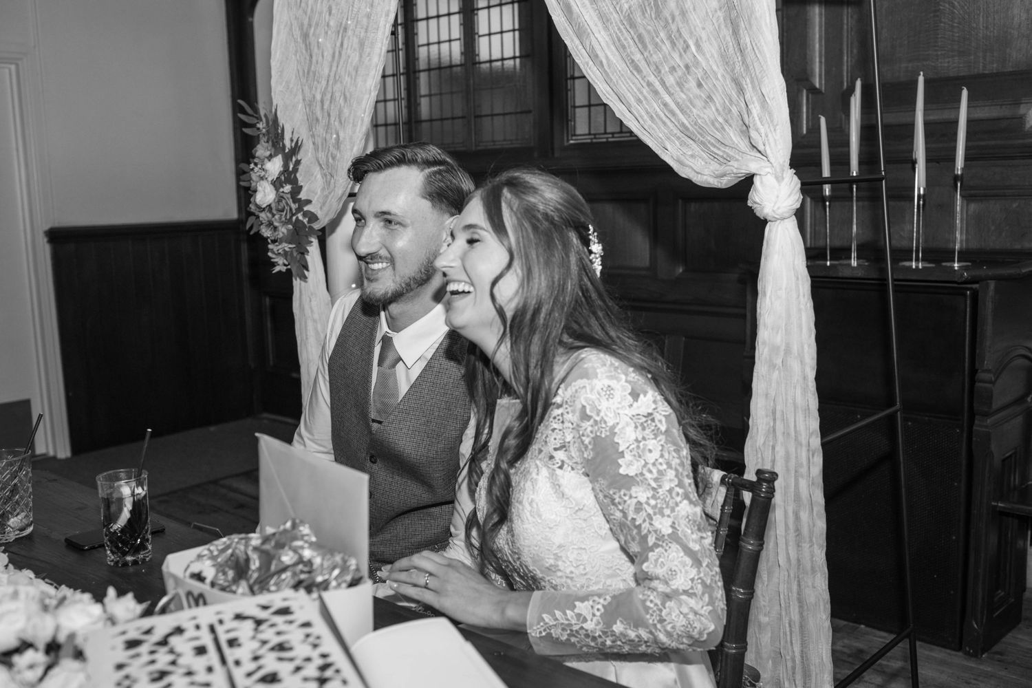 Black and white photo of a smiling bride and groom seated at a table during their wedding ceremony.