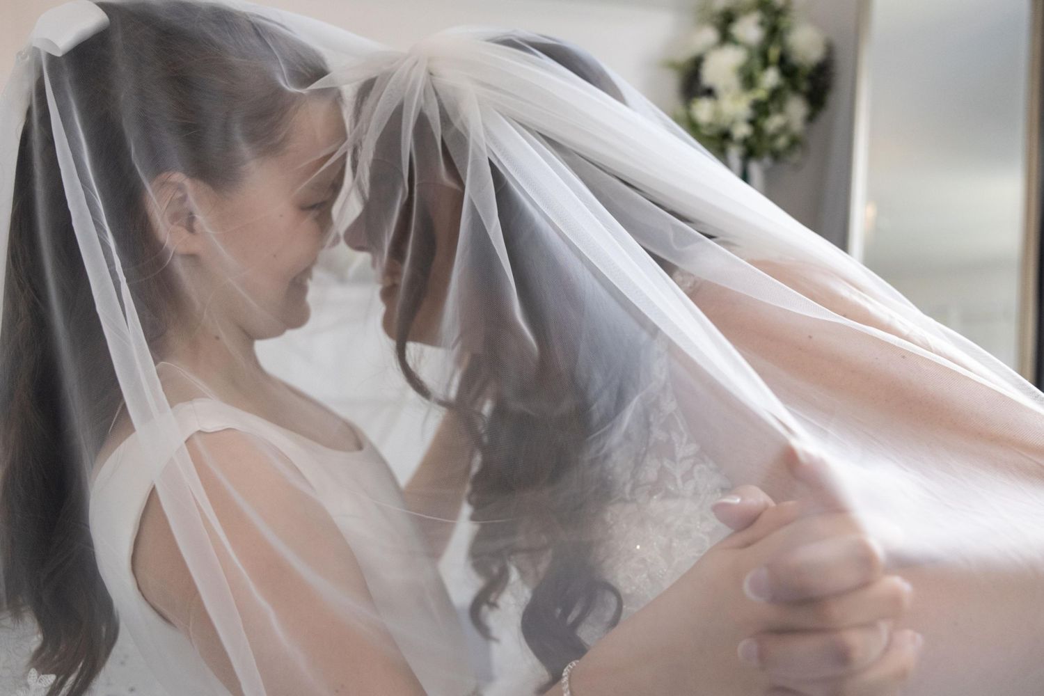 Two brides share an intimate moment under a flowing white wedding veil, holding hands in soft romantic lighting.