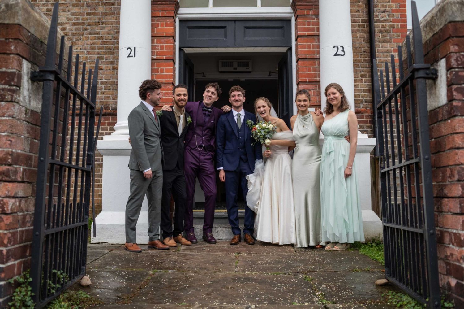 A joyful wedding party poses outside a brick building, with bride in white and guests in formal attire.