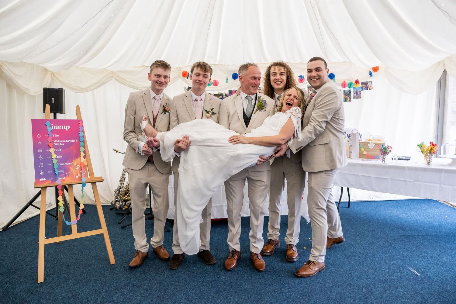 Five people in beige suits pose together in a marquee tent, laughing while holding a woman horizontally at a wedding.